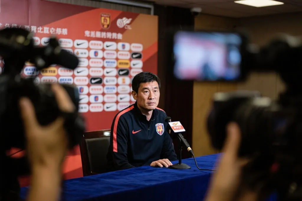 新闻发布会现场：灯光与相机对准教练，桌上话筒，紧张与克制